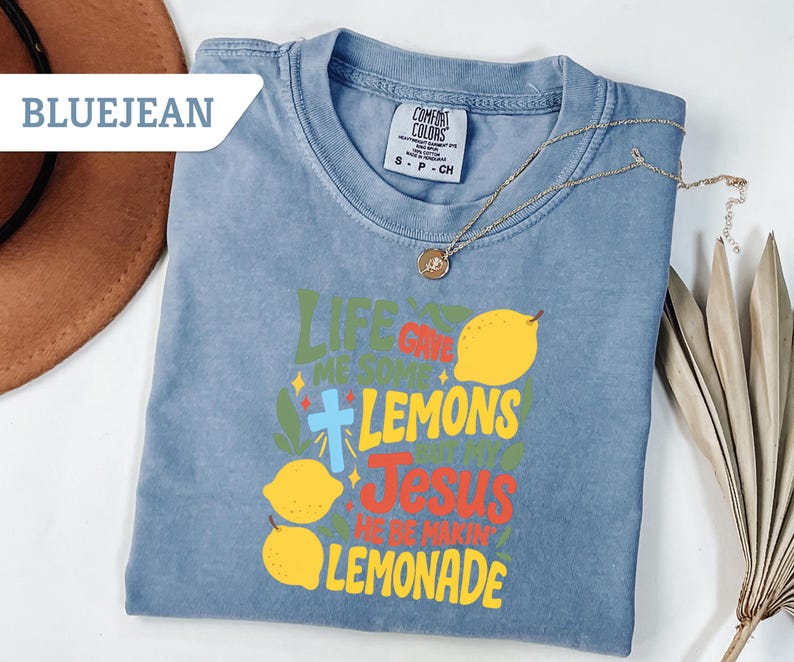 May include: Bluejean colored t-shirt with the text "Life Gave Me Some Lemons, So I Made Jesus Lemonade" in green, yellow, and red. The shirt features a cross and lemon graphics. A gold necklace and a brown hat are also visible.