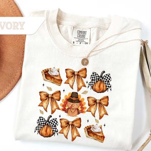 May include: Ivory-colored t-shirt featuring a Thanksgiving-themed design. The design includes illustrations of pumpkin pie slices, bows, pumpkins with checkered bows, and a turkey wearing a pilgrim hat. The shirt has a crew neck and short sleeves.