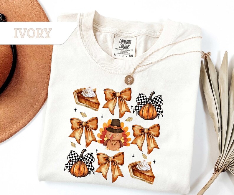 May include: Ivory-colored t-shirt featuring a Thanksgiving-themed design. The design includes illustrations of pumpkin pie slices, bows, pumpkins with checkered bows, and a turkey wearing a pilgrim hat. The shirt has a crew neck and short sleeves.
