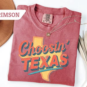 May include: Crimson-colored t-shirt with the phrase "Choosin' TEXAS" in a retro font, featuring the outline of Texas in yellow. A silver necklace with a pendant rests on the shirt. The shirt is folded on a white surface.