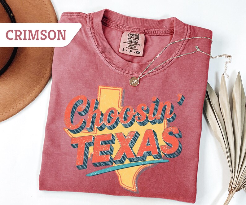 May include: Crimson-colored t-shirt with the phrase "Choosin' TEXAS" in a retro font, featuring the outline of Texas in yellow. A silver necklace with a pendant rests on the shirt. The shirt is folded on a white surface.