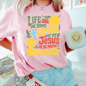 May include: A light pink t-shirt with the text "LIFE GAVE ME SOME LEMONS BUT MY JESUS HE BE MAKIN' LEMONADE" in various colors. The design includes lemons, a cross, and decorative elements. A white mug with a yellow smiley face is also visible.