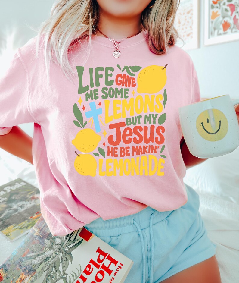 May include: A light pink t-shirt with the text "LIFE GAVE ME SOME LEMONS BUT MY JESUS HE BE MAKIN' LEMONADE" in various colors. The design includes lemons, a cross, and decorative elements. A white mug with a yellow smiley face is also visible.