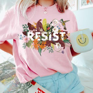 May include: A pink t-shirt with a floral design and the word "RESIST" printed on it. A hummingbird is perched on a flower in the design.