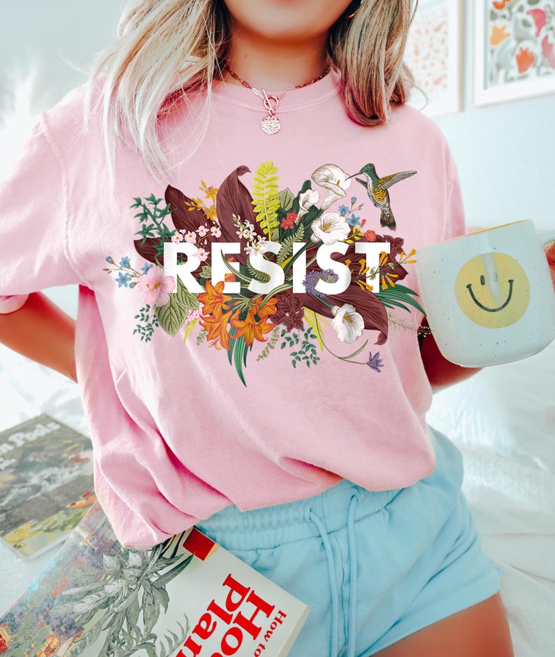May include: A pink t-shirt with a floral design and the word "RESIST" printed on it. A hummingbird is perched on a flower in the design.