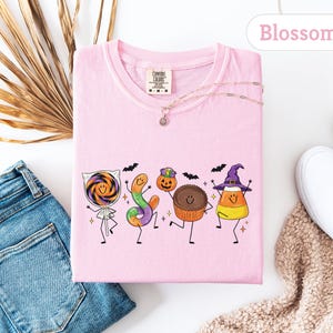 May include: A light pink t-shirt featuring a graphic of five anthropomorphic Halloween candies including a lollipop, a gummy worm, a pumpkin, a chocolate cup, and candy corn, all with smiling faces, thin black limbs, and small bats.