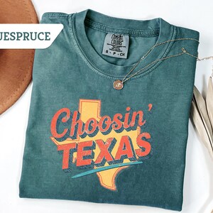 May include: Teal-colored t-shirt with the phrase "Choosin' TEXAS" in orange and red lettering, with a yellow Texas outline. A gold necklace with a pendant rests on the shirt. A brown hat and dried palm leaves are in the background.