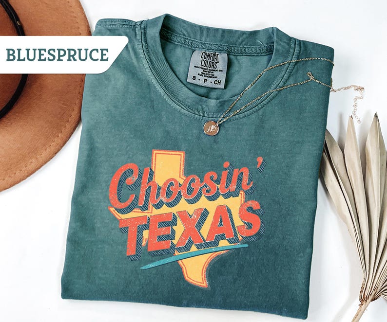 May include: Teal-colored t-shirt with the phrase "Choosin' TEXAS" in orange and red lettering, with a yellow Texas outline. A gold necklace with a pendant rests on the shirt. A brown hat and dried palm leaves are in the background.