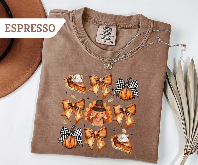 May include: Espresso-colored t-shirt with a fall-themed graphic. The design includes pumpkin pie slices, pumpkins with checkered bows, and a turkey wearing a pilgrim hat. A silver necklace is draped across the shirt.
