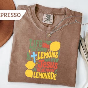 May include: Espresso-colored t-shirt with the text "Life gives you some lemons but my Jesus he be makin' lemonade" in green, yellow, red, and blue. The shirt features a cross and lemon graphics. A silver necklace with a pendant is on the shirt.
