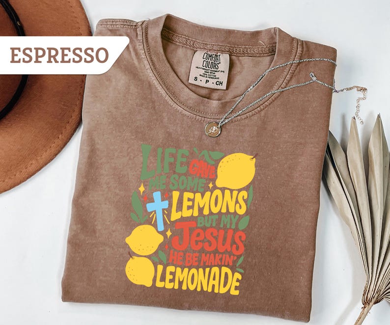 May include: Espresso-colored t-shirt with the text "Life gives you some lemons but my Jesus he be makin' lemonade" in green, yellow, red, and blue. The shirt features a cross and lemon graphics. A silver necklace with a pendant is on the shirt.