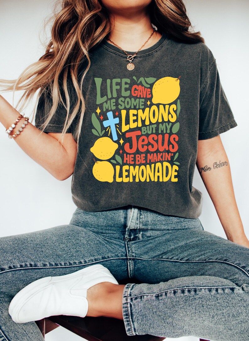 May include: Dark gray t-shirt with the text "Life Gave Me Some Lemons But My Jesus He Be Makin' Lemonade" in colorful lettering. The design includes lemons, a cross, and leaves. The shirt is paired with blue jeans and white sneakers.