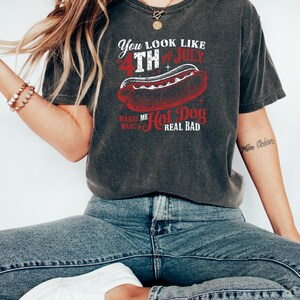 Op de afbeelding: Donkergrijs T-shirt met een afbeelding met de tekst "You look like 4th of July makes me want a hot dog real bad." De afbeelding toont een rode hotdog. De persoon draagt een blauwe spijkerbroek en witte sneakers.