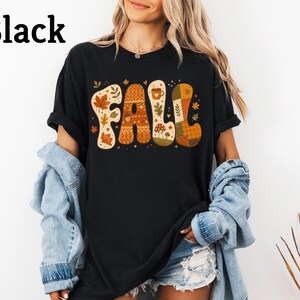 May include: A black t-shirt featuring the word FALL in large, stylized letters. Each letter contains autumn-themed patterns including maple leaves, acorns, mushrooms, and a coffee mug in shades of orange, brown, cream, and green. The text Black is visible.