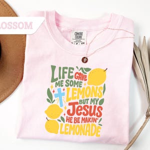 May include: A light pink t-shirt with the text "LIFE GAVE ME SOME LEMONS BUT MY JESUS HE BE MAKIN' LEMONADE" in green, yellow, and red. The shirt has a crew neck and short sleeves. A gold necklace with a pendant is on the shirt.