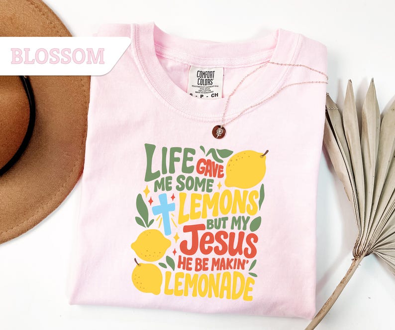 May include: A light pink t-shirt with the text "LIFE GAVE ME SOME LEMONS BUT MY JESUS HE BE MAKIN' LEMONADE" in green, yellow, and red. The shirt has a crew neck and short sleeves. A gold necklace with a pendant is on the shirt.