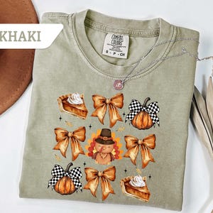 May include: Khaki-colored t-shirt featuring a fall-themed graphic. The design includes pumpkin pie slices, orange bows, pumpkins with checkered patterns, and a turkey wearing a pilgrim hat. A silver necklace is draped across the shirt.