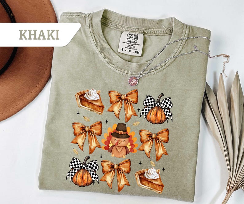 May include: Khaki-colored t-shirt featuring a fall-themed graphic. The design includes pumpkin pie slices, orange bows, pumpkins with checkered patterns, and a turkey wearing a pilgrim hat. A silver necklace is draped across the shirt.