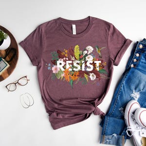 May include: A mauve t-shirt with a floral design and the word "RESIST" in white lettering.