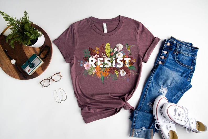 May include: A mauve t-shirt with a floral design and the word "RESIST" in white lettering.