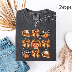 May include: Dark gray Comfort Colors t-shirt with a Thanksgiving-themed graphic. The design includes watercolor illustrations of bows, pumpkin pie, a turkey wearing a pilgrim hat, and pumpkins. A silver necklace is visible.
