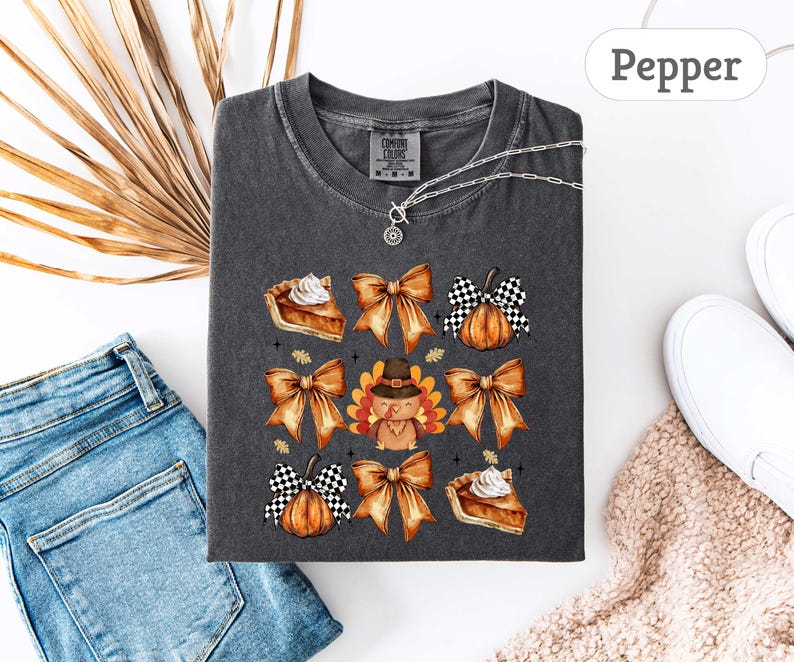 May include: Dark gray Comfort Colors t-shirt with a Thanksgiving-themed graphic. The design includes watercolor illustrations of bows, pumpkin pie, a turkey wearing a pilgrim hat, and pumpkins. A silver necklace is visible.