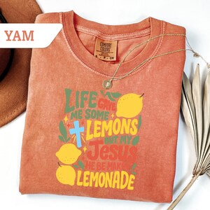 May include: A coral-colored t-shirt with the text "LIFE GAVE ME SOME LEMONS BUT MY JESUS HE BE MAKING LEMONADE" in green, yellow, and red. The design includes lemons and a blue cross. A gold necklace is draped across the shirt.