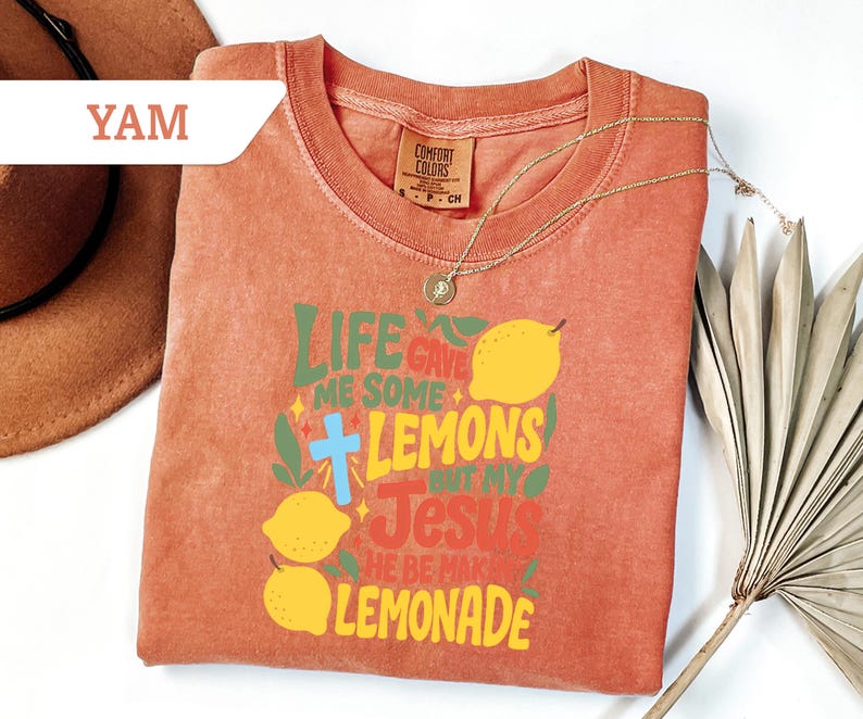 May include: A coral-colored t-shirt with the text "LIFE GAVE ME SOME LEMONS BUT MY JESUS HE BE MAKING LEMONADE" in green, yellow, and red. The design includes lemons and a blue cross. A gold necklace is draped across the shirt.
