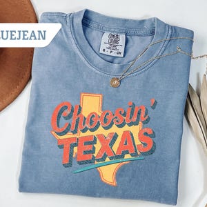 May include: Blue jean colored t-shirt with the words "Choosin' TEXAS" in red and orange lettering, with a yellow Texas outline. A gold necklace with a pendant is draped on the shirt. The shirt is folded on a white surface.