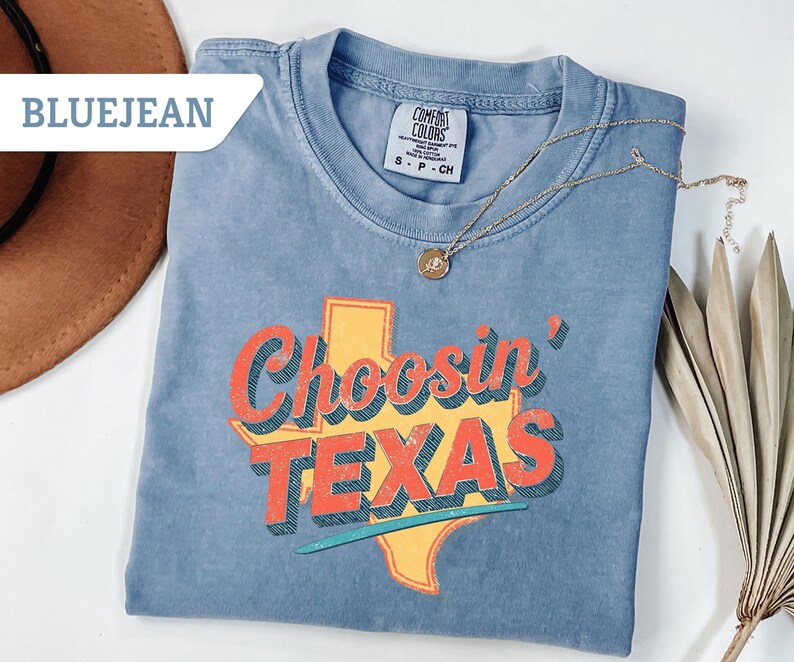 May include: Blue jean colored t-shirt with the words "Choosin' TEXAS" in red and orange lettering, with a yellow Texas outline. A gold necklace with a pendant is draped on the shirt. The shirt is folded on a white surface.