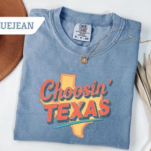 May include: Blue jean colored t-shirt with the text "Choosin' TEXAS" in orange and yellow. The graphic features the outline of Texas. A gold necklace and a brown hat are also visible.