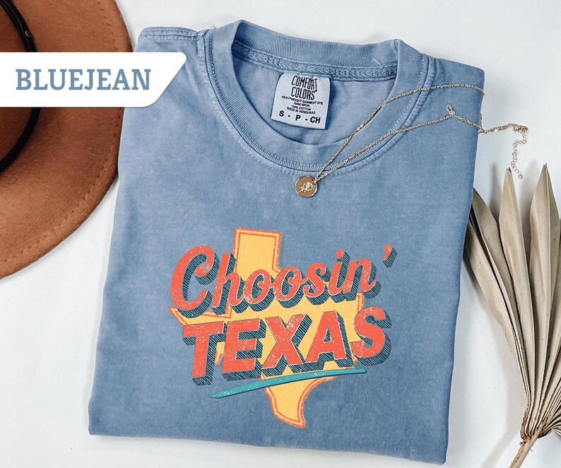 May include: Blue jean colored t-shirt with the text "Choosin' TEXAS" in orange and yellow. The graphic features the outline of Texas. A gold necklace and a brown hat are also visible.