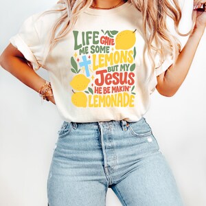 May include: Cream-colored t-shirt with the text "LIFE GAVE ME SOME LEMONS BUT MY JESUS HE BE MAKIN' LEMONADE" in green, blue, red, and yellow. The design includes lemons and a cross. The shirt is paired with light blue jeans.