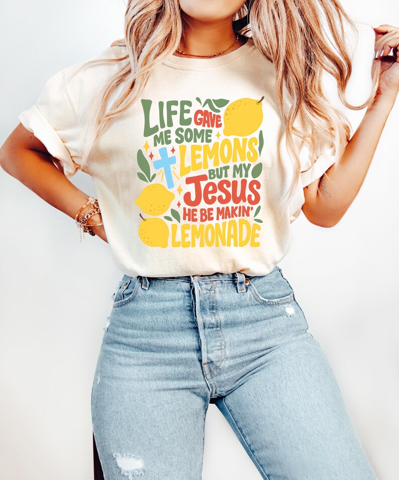 May include: Cream-colored t-shirt with the text "LIFE GAVE ME SOME LEMONS BUT MY JESUS HE BE MAKIN' LEMONADE" in green, blue, red, and yellow. The design includes lemons and a cross. The shirt is paired with light blue jeans.