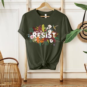 May include: A green t-shirt with a floral design and the word "RESIST" in white letters.