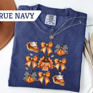 May include: True navy blue t-shirt with a Thanksgiving-themed graphic. The design includes images of pie slices, pumpkins with checkered patterns, bows, and a turkey wearing a hat. A gold necklace is draped across the shirt.