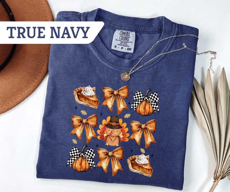 May include: True navy blue t-shirt with a Thanksgiving-themed graphic. The design includes images of pie slices, pumpkins with checkered patterns, bows, and a turkey wearing a hat. A gold necklace is draped across the shirt.