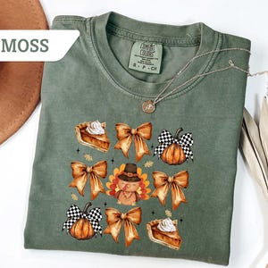 May include: Moss green t-shirt with a fall-themed graphic. The design includes illustrations of pie slices, bows, pumpkins, and a turkey wearing a hat. The shirt has a crew neck and short sleeves.