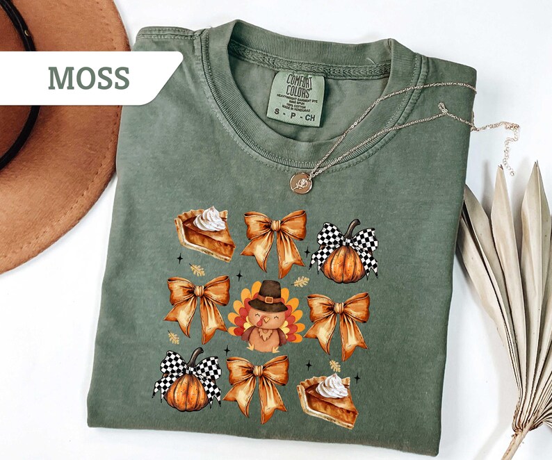 May include: Moss green t-shirt with a fall-themed graphic. The design includes illustrations of pie slices, bows, pumpkins, and a turkey wearing a hat. The shirt has a crew neck and short sleeves.