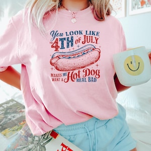 Op de afbeelding: Een lichtroze t-shirt met een afbeelding van een hotdog en de tekst: "You look like the 4th of July. Makes me want a hot dog real bad." Het shirt wordt gecombineerd met lichtblauwe shorts en een mok met een smiley.