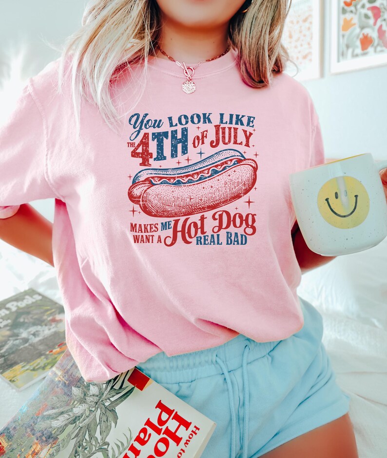 Op de afbeelding: Een lichtroze t-shirt met een afbeelding van een hotdog en de tekst: "You look like the 4th of July. Makes me want a hot dog real bad." Het shirt wordt gecombineerd met lichtblauwe shorts en een mok met een smiley.