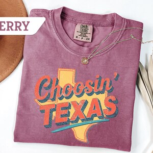 May include: Berry-colored t-shirt with the phrase "Choosin' TEXAS" in a retro font, featuring a yellow Texas outline. A gold necklace and a brown hat are also visible. The shirt is made of a soft material.