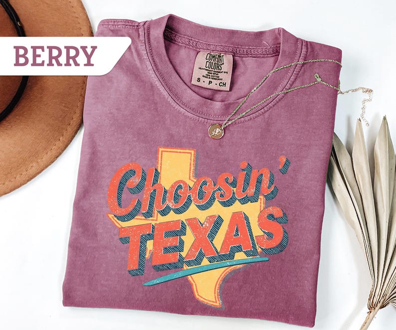 May include: Berry-colored t-shirt with the phrase "Choosin' TEXAS" in a retro font, featuring a yellow Texas outline. A gold necklace and a brown hat are also visible. The shirt is made of a soft material.
