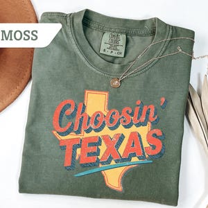 May include: A moss green t-shirt with the phrase "Choosin' TEXAS" in red and blue lettering over a yellow Texas outline. A gold necklace with a pendant rests on the shirt. A brown hat and dried plant are in the background.