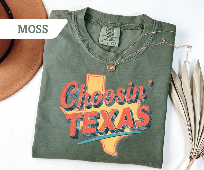 May include: A moss green t-shirt with the phrase "Choosin' TEXAS" in red and blue lettering over a yellow Texas outline. A gold necklace with a pendant rests on the shirt. A brown hat and dried plant are in the background.