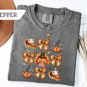 May include: A gray Comfort Colors t-shirt featuring a fall-themed design. The design includes images of pumpkin pie slices, orange bows, pumpkins with black and white checkered bows, and a turkey wearing a pilgrim hat.