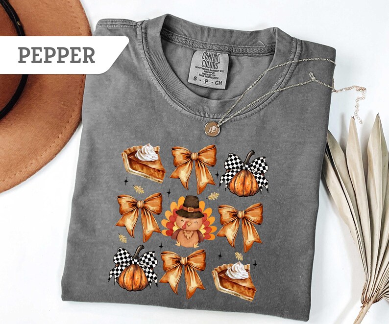 May include: A gray Comfort Colors t-shirt featuring a fall-themed design. The design includes images of pumpkin pie slices, orange bows, pumpkins with black and white checkered bows, and a turkey wearing a pilgrim hat.