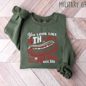 Op de afbeelding: Militairgroen sweatshirt met een afbeelding van een hotdog en de tekst "You look like 4th of July makes me want a hot dog real bad". Het ontwerp is in rood en wit.