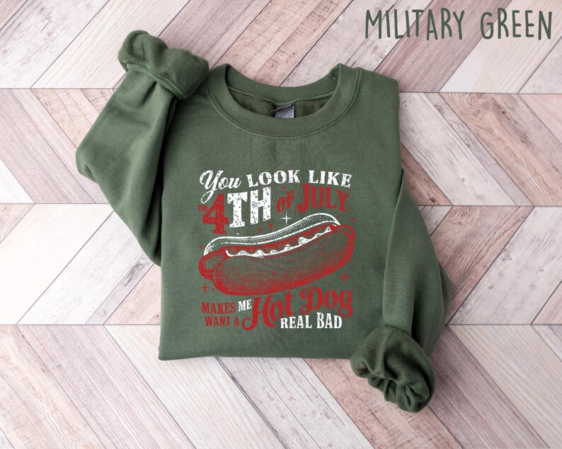 Op de afbeelding: Militairgroen sweatshirt met een afbeelding van een hotdog en de tekst "You look like 4th of July makes me want a hot dog real bad". Het ontwerp is in rood en wit.