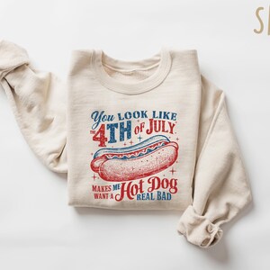 Op de afbeelding: Zandkleurige sweatshirt met een afbeelding van een hotdog en de tekst "You look like the 4th of July makes me want a hot dog real bad" in rood en blauw. De sweatshirt is opgevouwen.